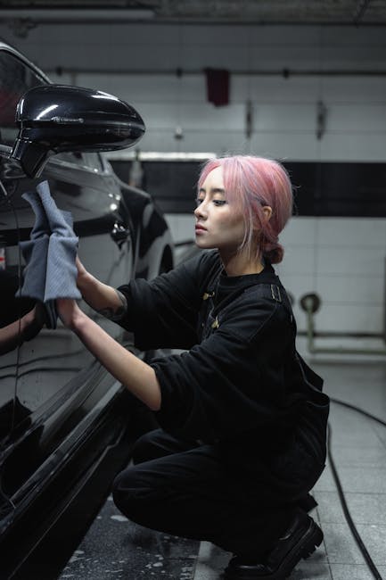 A woman with pink hair cleaning a black car in an indoor carwash setting, focused and skilled.